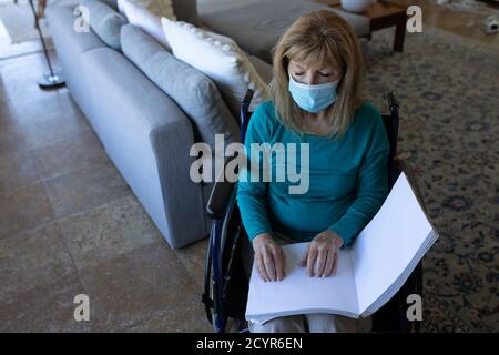 Donna caucasica anziana che passa il tempo a casa, seduto su sedia a rotelle e leggendo un libro con le mani, indossando la maschera facciale. Distanza sociale durante il Cov Foto Stock