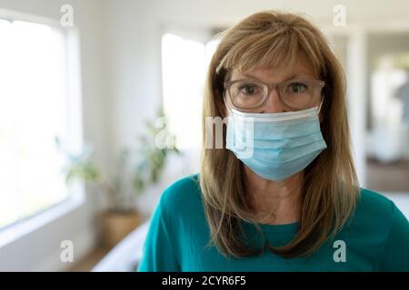 Ritratto di una donna caucasica anziana che trascorre del tempo a casa, in piedi nel suo soggiorno indossando una maschera facciale. Distanza sociale durante Covid 19 Coronav Foto Stock