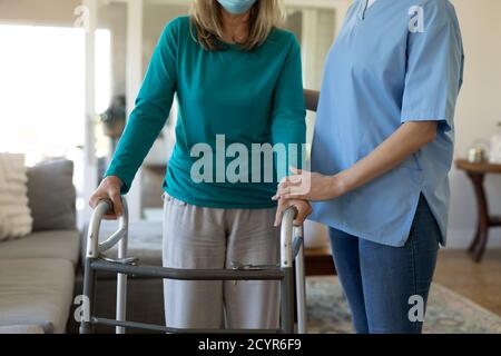 Donna caucasica anziana a casa visitata da infermiere caucasica, camminando con un camminatore, indossando la maschera facciale. Assistenza medica a casa durante Covid 19 Coro Foto Stock