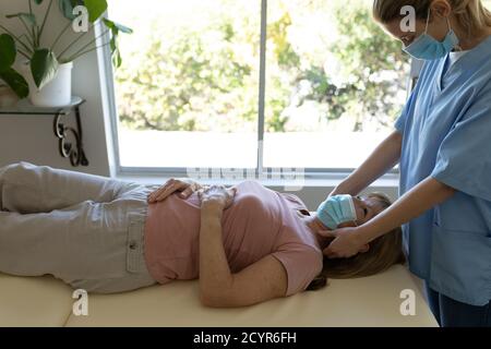 Donna caucasica anziana a casa visitata da infermiere caucasica, massaggiando il collo, indossando maschere facciali. Assistenza medica a casa durante Covid 19 Coronav Foto Stock