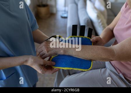 Donna caucasica senior a casa visitata da un'infermiera caucasica, indossando un supporto per il polso. Assistenza medica a casa durante il Covid 19 Coronavirus quarant Foto Stock