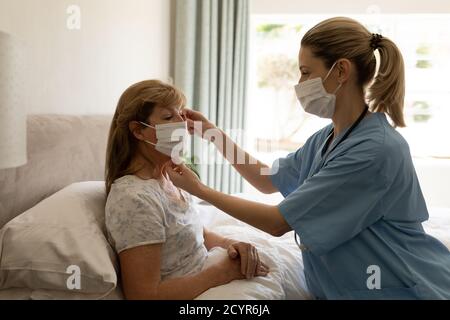 Donna caucasica anziana a casa visitata da infermiere caucasica, mettendo una maschera sul viso, infermiera che indossa la maschera. Assistenza medica a casa durante Covid 1 Foto Stock