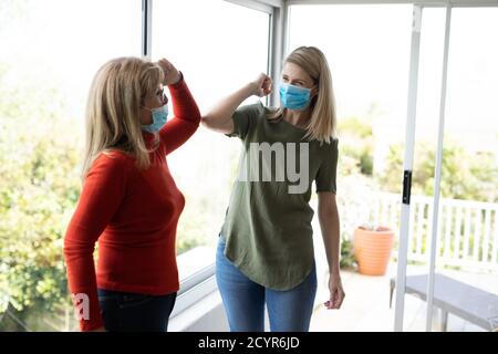 Donna caucasica anziana e sua figlia adulta a casa, indossando maschere facciali e salutarsi a vicenda toccando gomiti. Distanza sociale, salute e hy Foto Stock