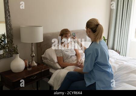 Donna caucasica anziana a casa visitata da infermiere caucasica, controllo della temperatura. Assistenza medica a casa durante la quarantena di Coronavirus Covid 19. Foto Stock