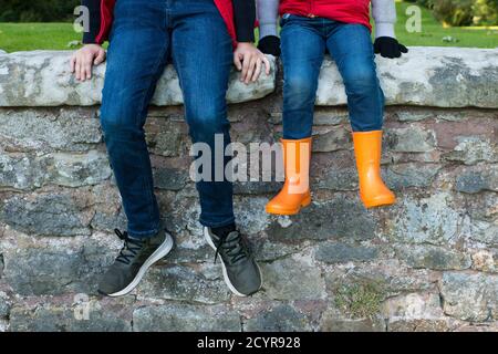 primo piano dei piedi e delle gambe dei bambini che indossano scarpe da ginnastica e stivali wellington dai colori vivaci, seduti su un muro di pietra all'aperto sotto il sole d'autunno Foto Stock