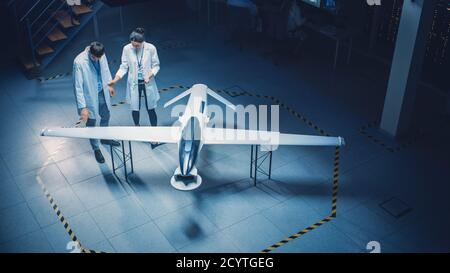 Due ingegneri aerospaziali lavorano sul prototipo di drone di un veicolo aereo senza equipaggio. Scienziati dell'aviazione in White Coats che parlano, utilizzando Tablet computer. Industriale Foto Stock