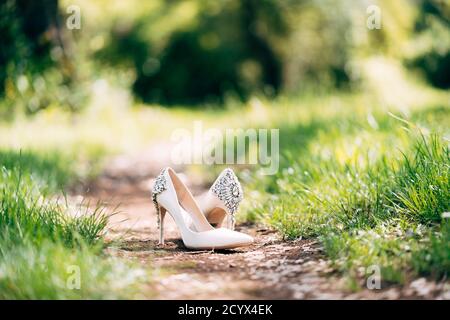 Scarpe da sposa bianche da donna della sposa, con tacchi alti, decorate con pietre di gioielli. Su un sentiero forestale tra l'erba verde. Foto Stock