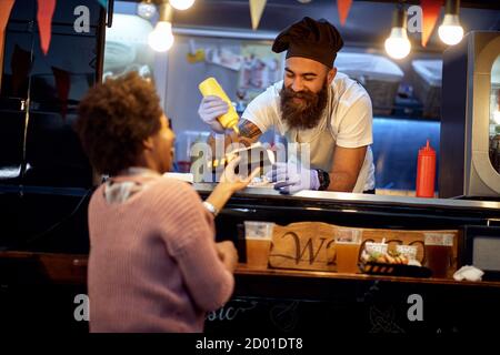 Food Truck Employee consegna un Sanwich appena fatto a. Una giovane donna afro-americana felice Foto Stock