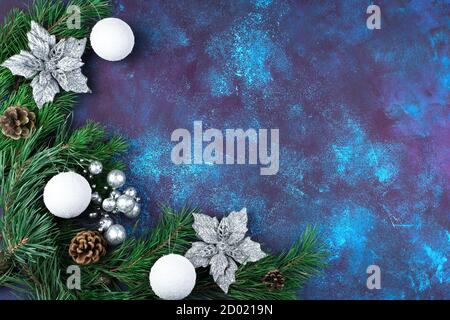 Rami di pino verde sono decorati con bellissimi fiori d'argento, coni e giocattoli di Natale su uno sfondo blu di cemento. Foto Stock
