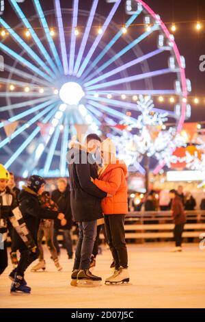 Uomo e donna innamorati in abbraccio su arena di ghiaccio contro di ruota Ferris in serata di Natale. Coppia a pista di pattinaggio, romanticismo e incandescente città Foto Stock