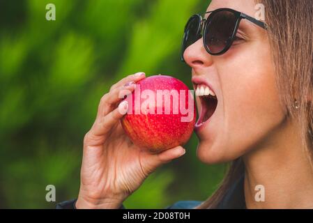 Carino donna vegana sana in occhiali da sole neri che mordicchiano la mela succosa rossa. Foto Stock