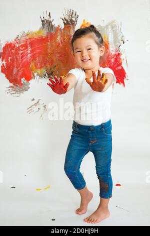 Bambina asiatica in t-shirt bianca con mani cosparse di vernice sullo sfondo di un muro dipinto. Foto Stock