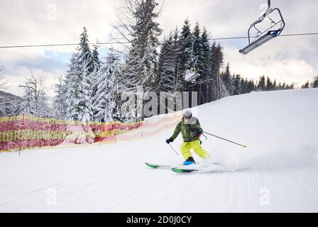Sciatori maschili e carving girare su pista da sci preparata presso la stazione sciistica dopo aver preso la seggiovia sulla montagna innevata. Discesa di sci di sicurezza in alta montagna. Seggiovia su collina boscosa sullo sfondo Foto Stock