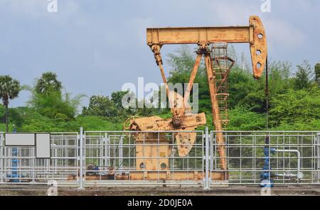 Vecchio martinetto a pompa dell'olio arrugginito che estrae olio grezzo e naturale gas da un pozzo di petrolio in fondo di campo di petrolio verde Foto Stock