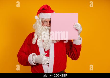 Attore maschio in un vestito di Babbo Natale tiene dentro le sue mani un foglio rosa di carta per i dischi e. posa su sfondo giallo Foto Stock