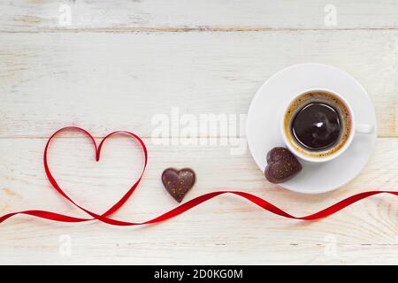 Tazza di caffè americano nero con cioccolato dorato a forma di cuore e nastro di raso rosso su fondo di legno. Vista dall'alto. Foto Stock