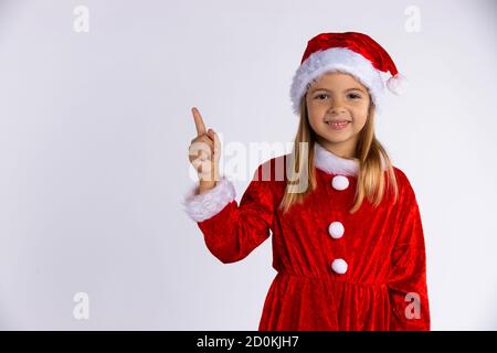 Piccola ragazza in cappello di Santa e costume con bel sorriso punta il dito. Isolato su sfondo bianco Foto Stock