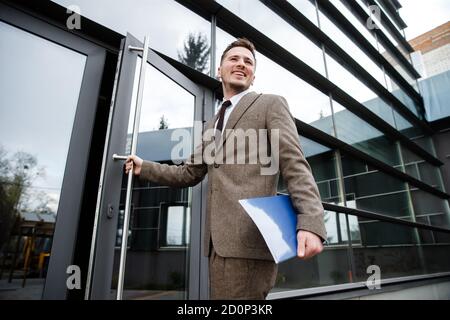 Giovane lavoratore d'ufficio che entra nell'edificio con una cartella in mano. Sta sorridendo. Foto Stock
