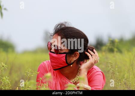 Una giovane donna di stile indiana che si pone in un campo con una maschera facciale rossa per prevenire la malattia della corona, concetto per proteggere gli agricoltori, lavoratori, operaio all'aperto Foto Stock