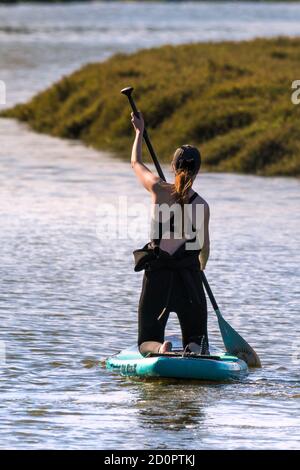 Vista posteriore di una vacanziera femminile inginocchiata su una tavola Stand Up in stile Sandbanks sul fiume Gannel a Newquay in Cornovaglia. Foto Stock