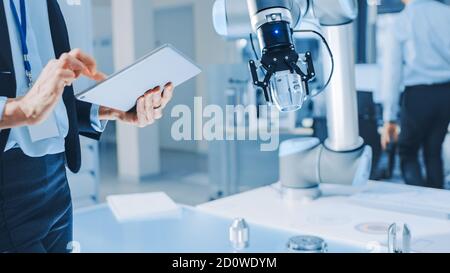 L'elegante ingegnere di robotica industriale femminile utilizza un Tablet computer per programmare e manipolare il braccio del robot per sollevare e spostare oggetti. Ricerca tecnologica Foto Stock