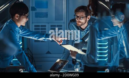 Team di ingegneri con notebook e tablet analizza e discute di come funziona un braccio robotico futuristico e muove un oggetto metallico. Sono in alto Foto Stock