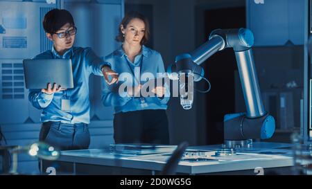 Uomo e donna ingegneri con notebook e tablet analizzare e discutere come un braccio robotico futuristico funziona e muove un oggetto metallico. Sono in alto Foto Stock