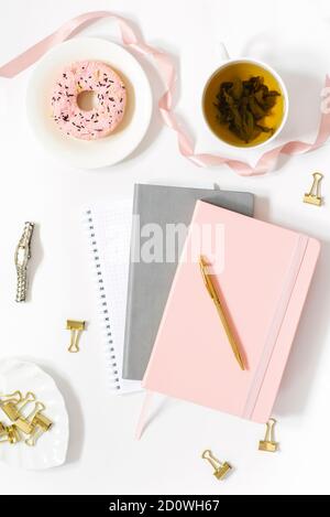 Piatto per un blogger di moda o freelancer. Diario rosa e grigio, tazza di tè verde, ciambella, orologio da polso e clip su uno sfondo bianco. Foto Stock