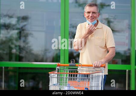 L'uomo sorridente maturo stende una maschera mentre se ne esce un supermercato con un carrello - focus su faccia Foto Stock