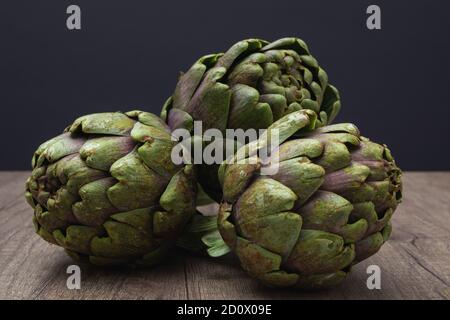 Tre carciofi viola freschi con gambo su un rustico superficie in legno e sfondo scuro Foto Stock