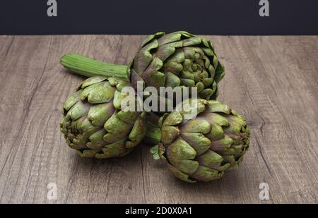 Tre carciofi viola freschi con gambo su un rustico superficie in legno e sfondo scuro Foto Stock