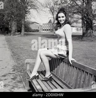 Nei terreni di un college londinese, una giovane donna attraente, forse una studentessa o forse un modello, seduta su una panchina di legno in una parte superiore a manica corta e gonna corta, Londra, Inghilterra, Regno Unito, all'inizio degli anni '70. Foto Stock