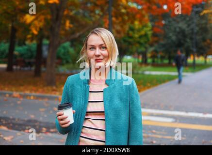 Donna caucasica sorridente bionda con tazzina di caffè da asporto che cammina nel parco autunnale. Attraente donna in piedi su crocevia Foto Stock