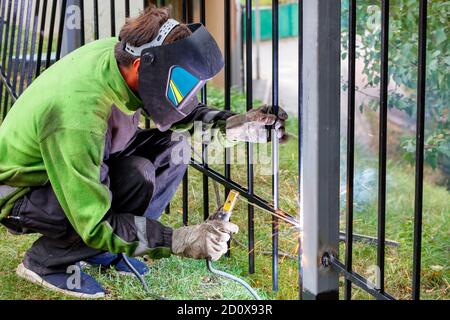 Una saldatrice che indossa indumenti protettivi, casco e guanti salda una recinzione metallica intorno all'area di lavoro. Foto Stock