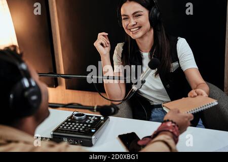 Portait di felice radio host femminile ridendo mentre si parla con ospite maschile, presentatore e dando un notebook a lui, moderando uno spettacolo dal vivo in studio Foto Stock