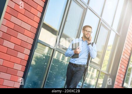 Giovane uomo d'affari caucasico felice che indossa camicia blu e occhiali tenendo il suo laptop, parlando al telefono e sorridendo mentre cammina all'aperto Foto Stock