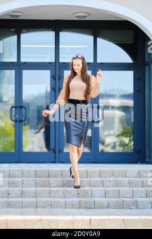 Donna giovane positiva in blusa e gonna di cuoio che scende il scale vicino all'edificio degli uffici Foto Stock