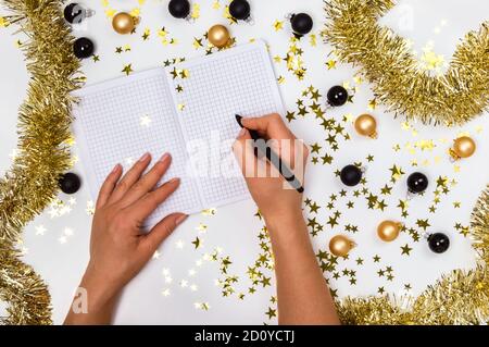 Mani femminili con penna e taccuino tra baubles natalizi dorati e neri, stelle garland e confetti. Lista dei desideri, risoluzioni di Capodanno o piano 2021 Foto Stock