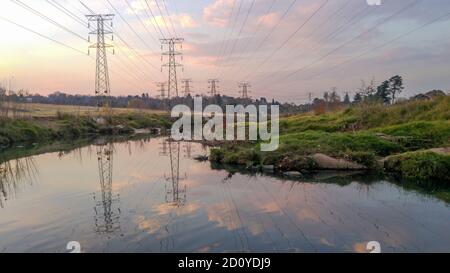 Una bella scena di erba verde, alberi e acqua fluente viziata dalla marcia di linee elettriche ad alta tensione attraverso il centro. Foto Stock