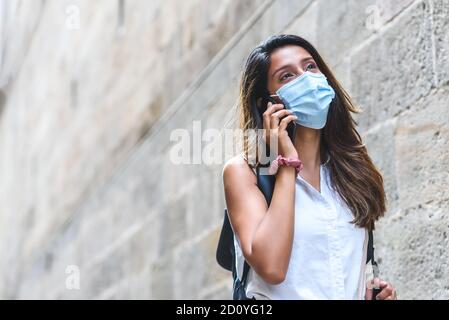 giovane donna indiana che indossa una maschera che parla al telefono in strada vicino a un muro con grande spazio di copia a sinistra Foto Stock