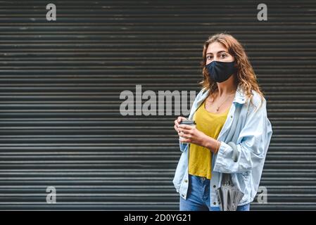 giovane donna caucasica rossa con una maschera nera che rimane sulla strada con una tazza di carta di take away caffè con sfondo nero Foto Stock