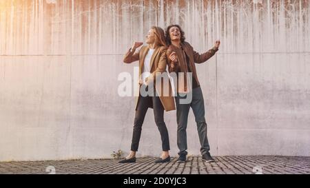 Cheerful Girl e Happy Young Man with Long Hair stanno ballando attivamente su una strada accanto a un muro urbano di cemento. Indossano giacca in pelle marrone e. Foto Stock