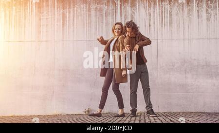 Cheerful Girl e Happy Young Man with Long Hair stanno ballando attivamente su una strada accanto a un muro urbano di cemento. Indossano giacca in pelle marrone e. Foto Stock