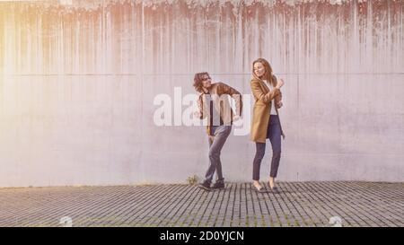 Cheerful Girl e Happy Young Man con i capelli lunghi stanno ballando attivamente su una strada mentre un uomo d'affari e un ragazzo con uno smartphone Pass by. Indossano Foto Stock