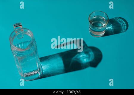 Un bicchiere con acqua accanto a una caraffa su un sfondo blu chiaro in cui vengono proiettate le ombre Foto Stock