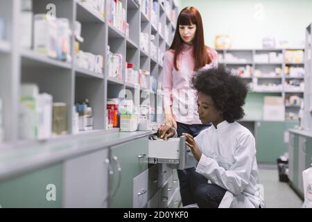 Donna sorridente piuttosto giovane, comprando le medicine alla farmacia., mentre la donna africana farmacista aiutandola e mostrando alcune droghe nel cassetto. Femmina Foto Stock