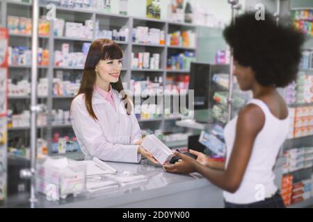 Giovane bella donna caucasica farmacista aiutare una donna africana cliente, dispensando la sua medicina di prescrizione con un sorriso amichevole. Farmacista e. Foto Stock