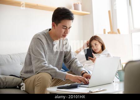 giovane coppia asiatica che soggiorna a casa con il marito che lavora usando computer portatile e moglie lettura ebook Foto Stock