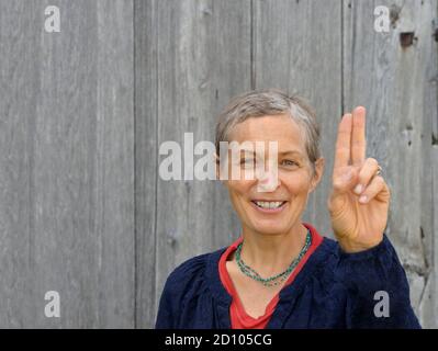 Donna caucasica positiva di mezza età con capelli corti (homesteader) prende un giuramento con due dita, di fronte ad un vecchio fondo di legno di fienile. Foto Stock