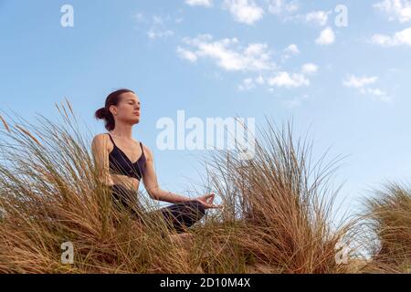 Donna matura meditando sulla spiaggia nelle dune di sabbia con uno sfondo blu Foto Stock